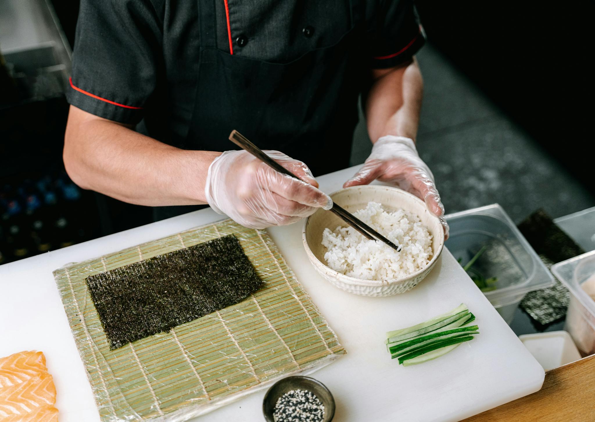 Close-up of a chef making sushi with chopsticks, rice, and nori on a cutting board.